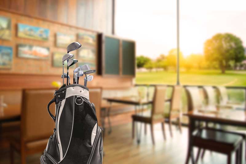 golf course loans and financing - Photo description: golf clubs in a golf bag standing upright in a golf course club house restaurants, which is out of focus behind it.