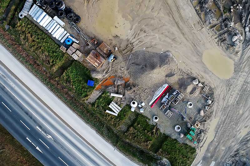 Construction site from above, revealing excavated soil, building materials, and active development next to a flowing highway